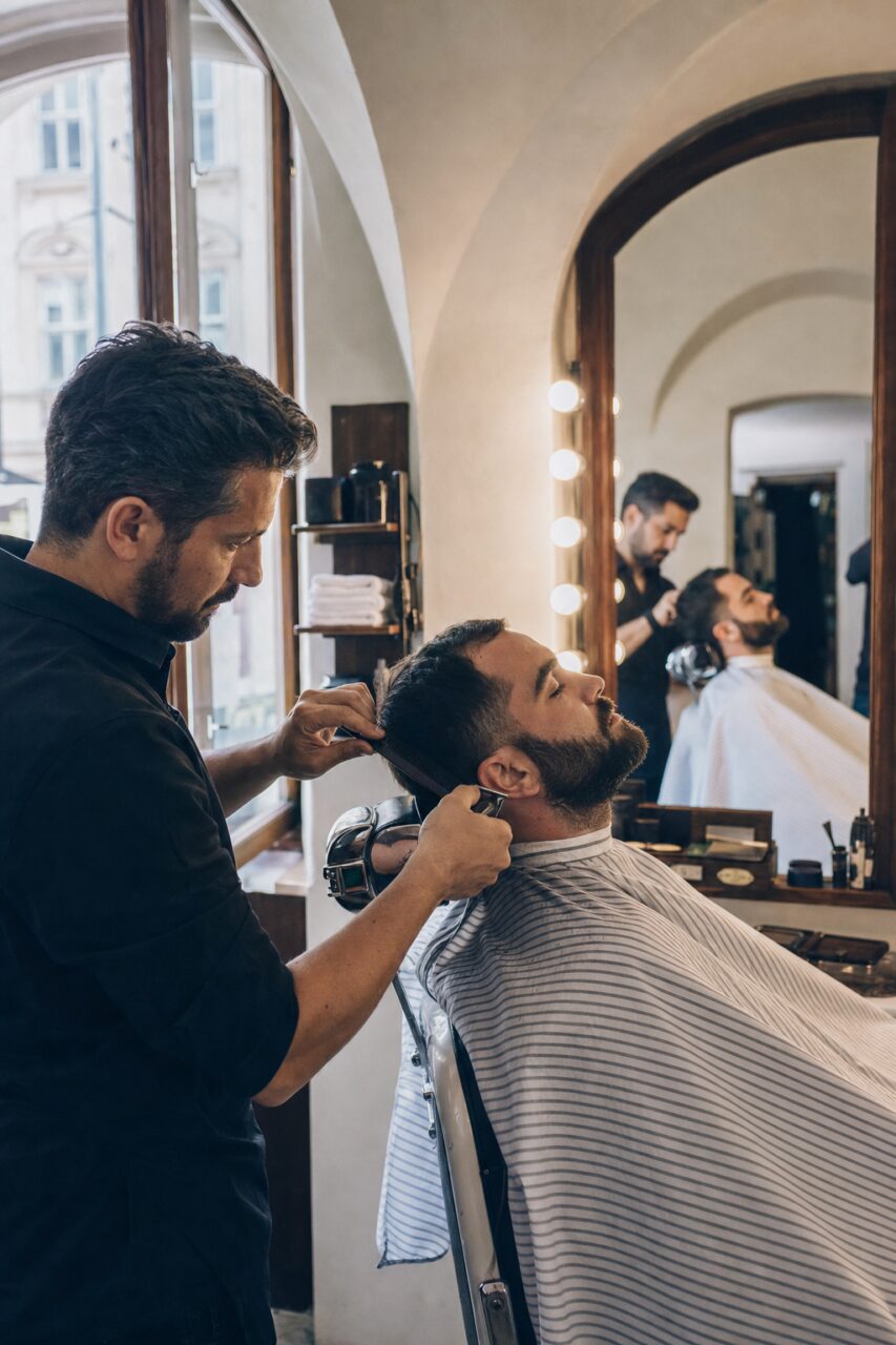 Barbiere usa la tosatrice sulla barba di un cliente sotto una mantella a righe davanti a uno specchio illuminato