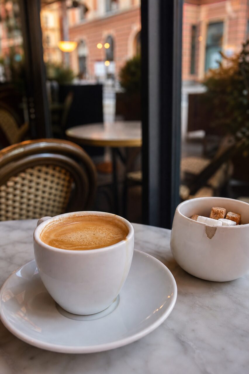 Café com leite em chávena branca junto de um açucareiro perto da janela