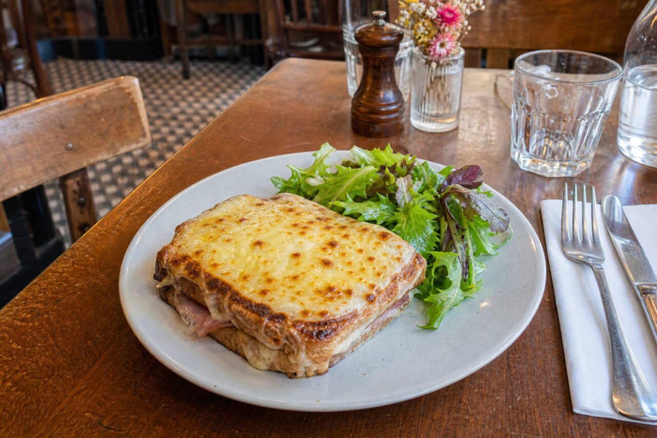 Gegratineerde croque monsieur met salade, glazen en bestek op een brasserietafel