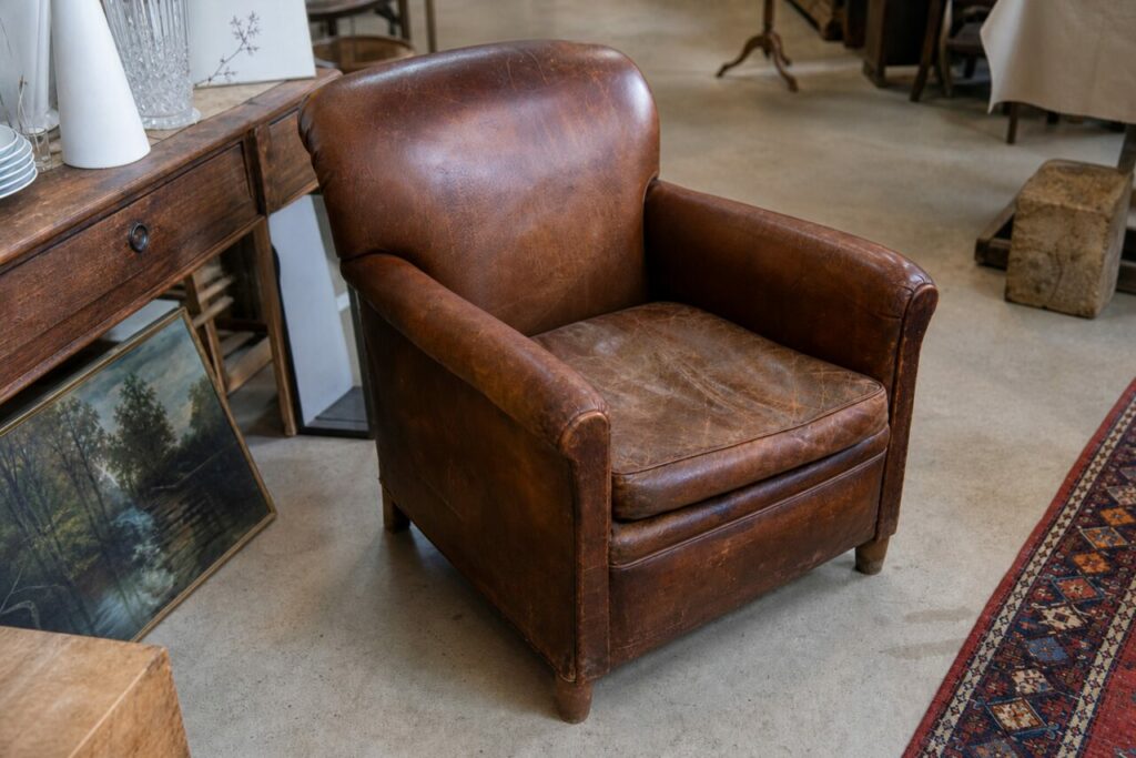 Fauteuil club en cuir brun vu en légère plongée au milieu du décor d’antiquaire