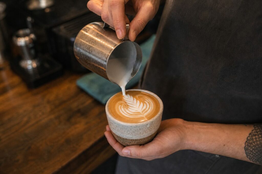 Hand gießt Milch in eine Kaffeetasse und vollendet ein Blattmuster an der Theke
