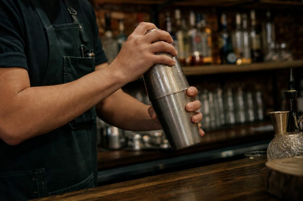 Hände halten einen Metallshaker vor den Flaschen einer Bar