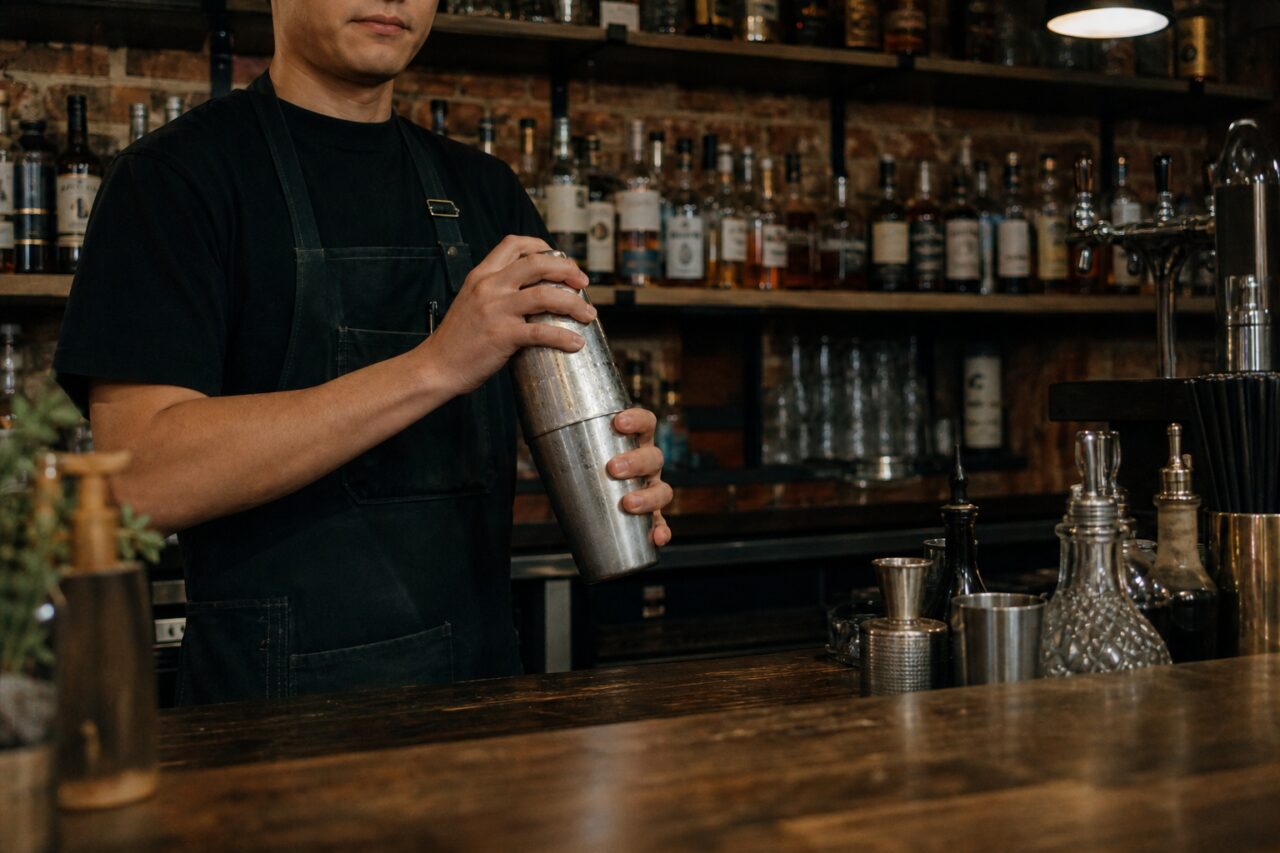 Person in einer Schürze bereitet mit einem Shaker einen Cocktail an der Bartheke zu