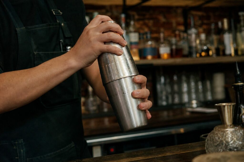 Person in einer Schürze schüttelt einen Metallshaker hinter der Bartheke