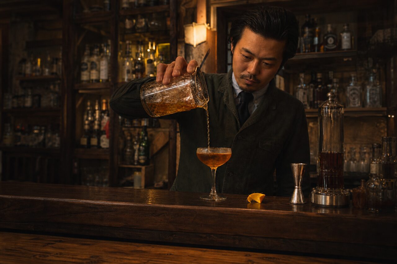Een amberkleurige drank wordt in een coupglas geschonken met de bar op de achtergrond