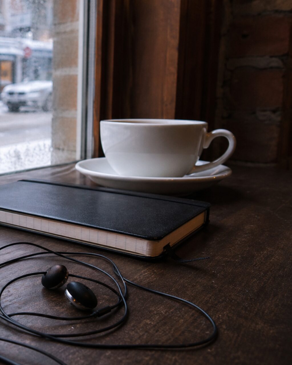 Tasse, Notizbuch und Ohrhörer auf einem Tisch am Fenster